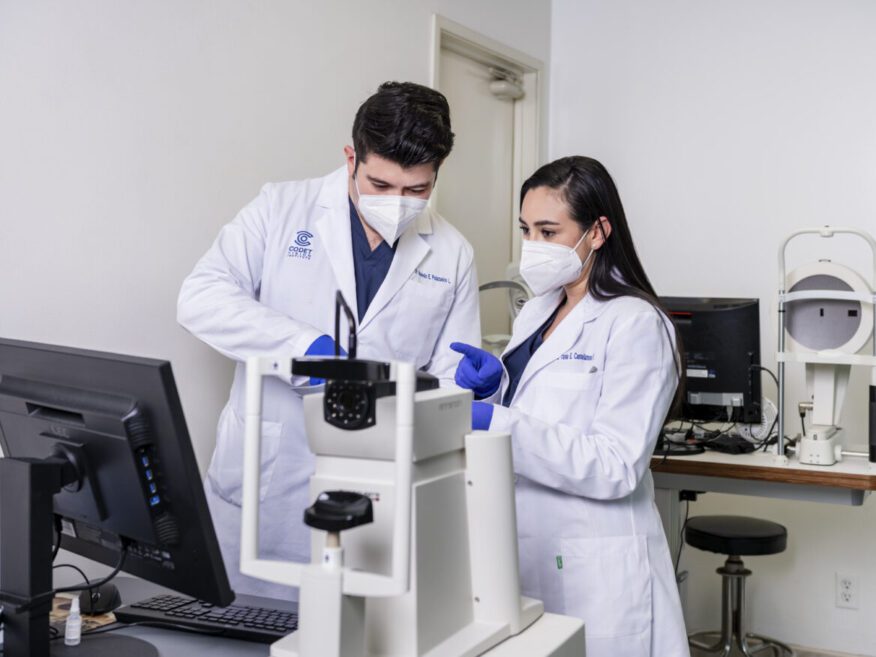Two medical professionals in lab coats and masks operating diagnostic eye equipment in a clinic