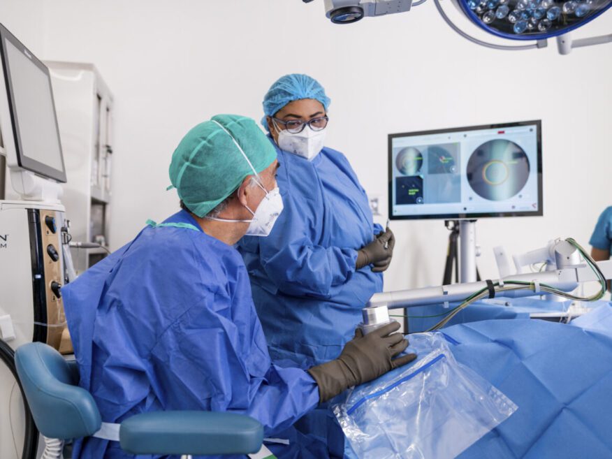 Two surgeons in protective gowns and masks are preparing for an eye procedure with medical monitors in the background.