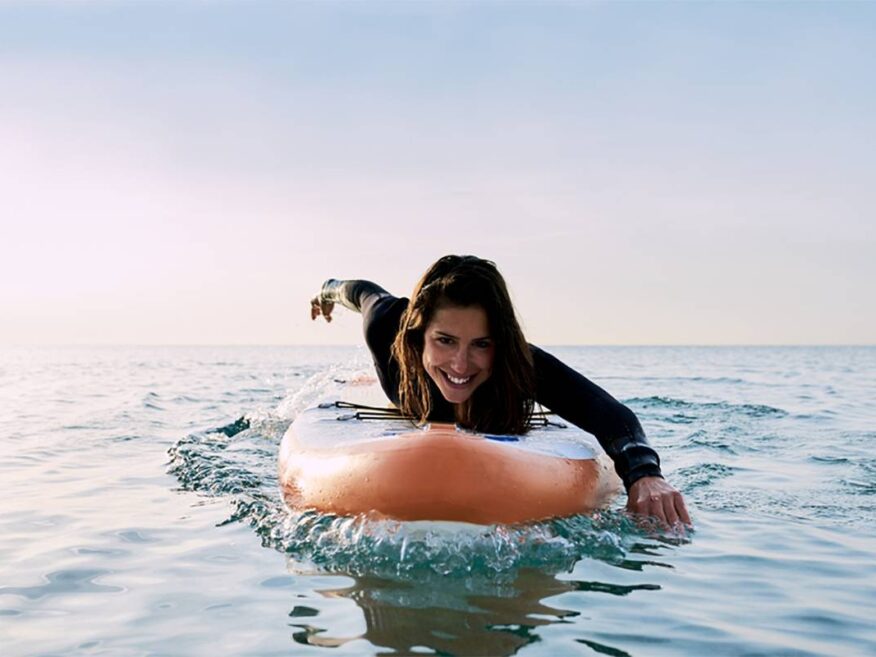 Smiling woman paddling on a surfboard in the ocean during sunset.